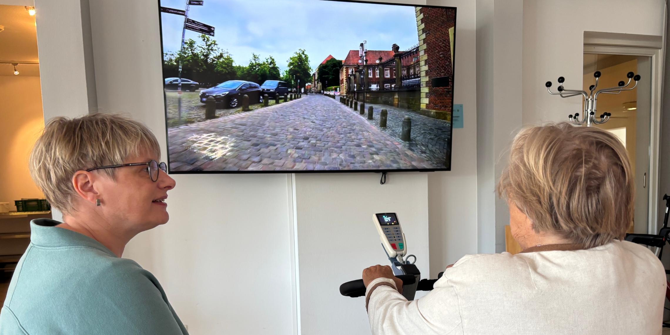 Eine Seniorin radelt auf einem Heimtrainer und verfolgt auf einem Bildschirm ihre virtuelle Radtour durch Münster, beobachtet von einer Mitarbeiterin des Sozialen Dienstes.