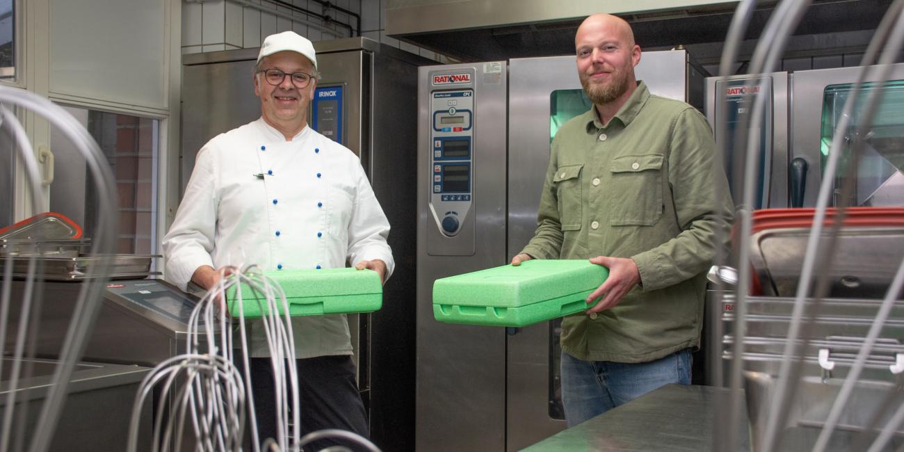 Küchenleiter Markus Lütjann und Einrichtungsleiter Sebastian Stödtke stehen in der Großküche des Henricus-Stifts und zeigen Warmhaltebehälter für das Essen auf Rädern.