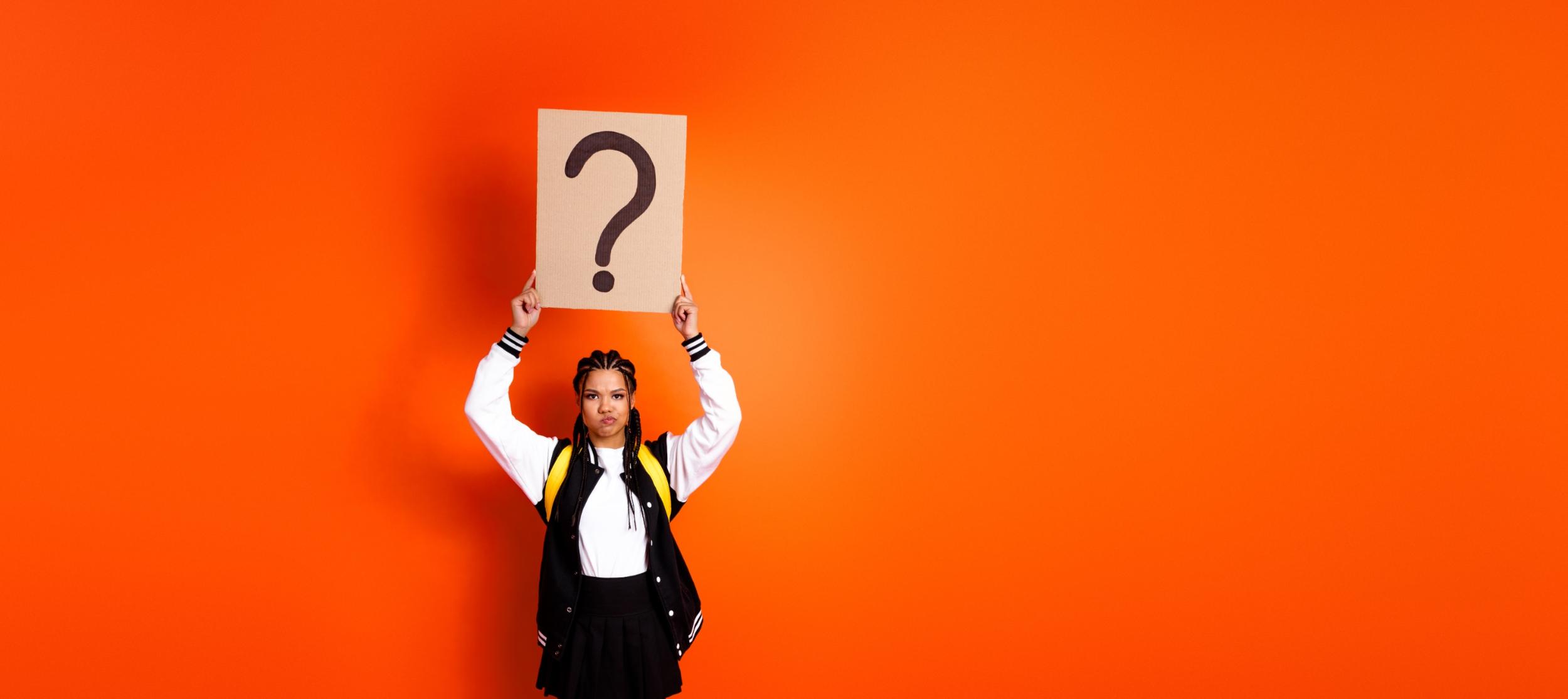 Eine junge Frau hält mit beiden Händen ein Plakat hoch, auf dem ein Fragezeichen steht.